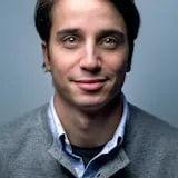 Professional headshot of a smiling man with dark hair wearing a gray sweater over a blue collared shirt