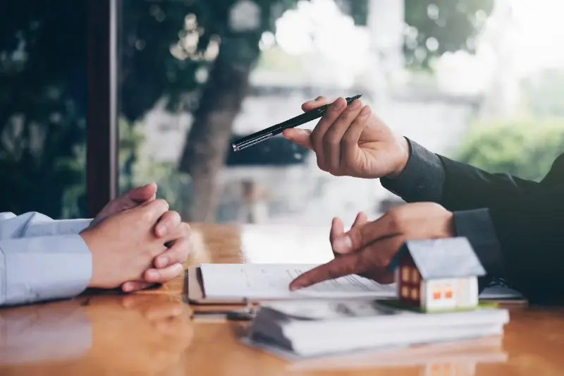 Real estate agent offering pen to client at wooden desk with property documents and model house