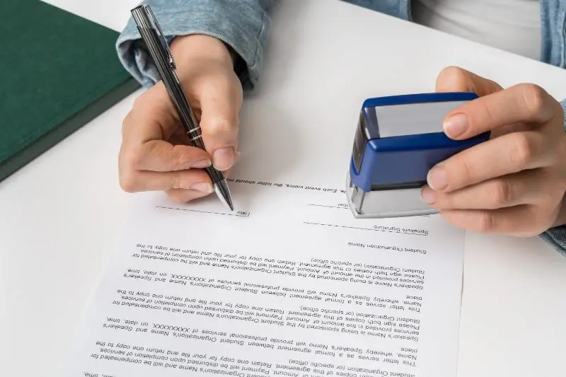 Person signing a document with a pen while holding a blue stamp
